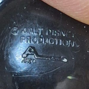 Puede incluir: Primer plano de un objeto de pl&aacute;stico negro con el texto "&copy; WALT DISNEY PRODUCTIONS" y el logotipo "APCO". El objeto parece ser un objeto de colecci&oacute;n vintage, posiblemente un juguete o una figura.