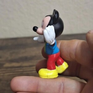 May include: A small Mickey Mouse figurine. The figure has a black head and ears, a red pair of pants, yellow shoes, a blue shirt, and white gloves. The figurine is in a pose with one arm raised.
