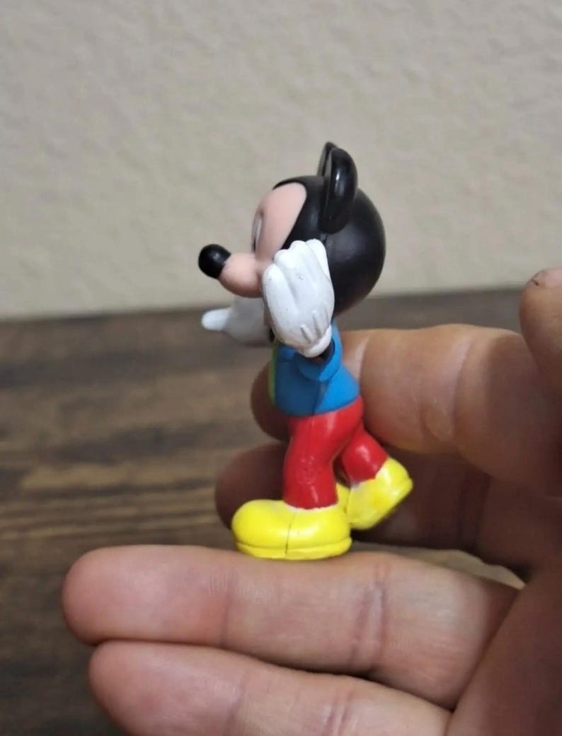 May include: A small Mickey Mouse figurine. The figure has a black head and ears, a red pair of pants, yellow shoes, a blue shirt, and white gloves. The figurine is in a pose with one arm raised.
