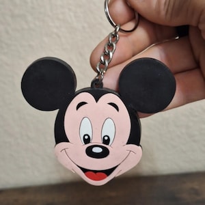 May include: A Mickey Mouse keychain featuring a smiling face with black ears, a pink face, and a red tongue. The keychain has a silver chain and ring. The design is a classic cartoon character.