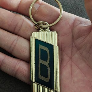 May include: Gold-toned metal keychain with a rectangular pendant. The pendant features a dark teal-colored inset with a capital letter "B" in a lighter gold color. The keychain has a ring for attaching keys.