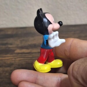 May include: A small Mickey Mouse figurine. The figure has a black head and ears, a pink face, a blue shirt, red pants, yellow shoes, and white gloves. The figurine is held in a hand.