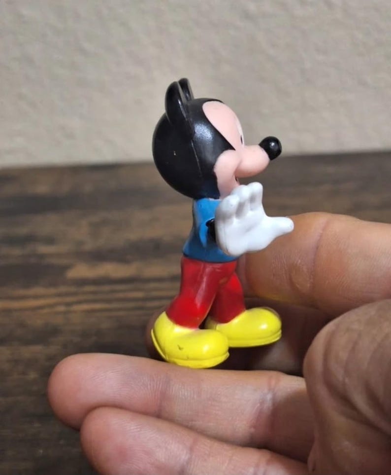 May include: A small Mickey Mouse figurine. The figure has a black head and ears, a pink face, a blue shirt, red pants, yellow shoes, and white gloves. The figurine is held in a hand.