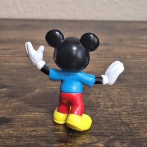 May include: A small Mickey Mouse figurine, viewed from the back, with arms outstretched. The figure has a black head with large ears, a blue shirt, red pants, yellow shoes, and white gloves. The word "DISNEY" is printed on the shirt.