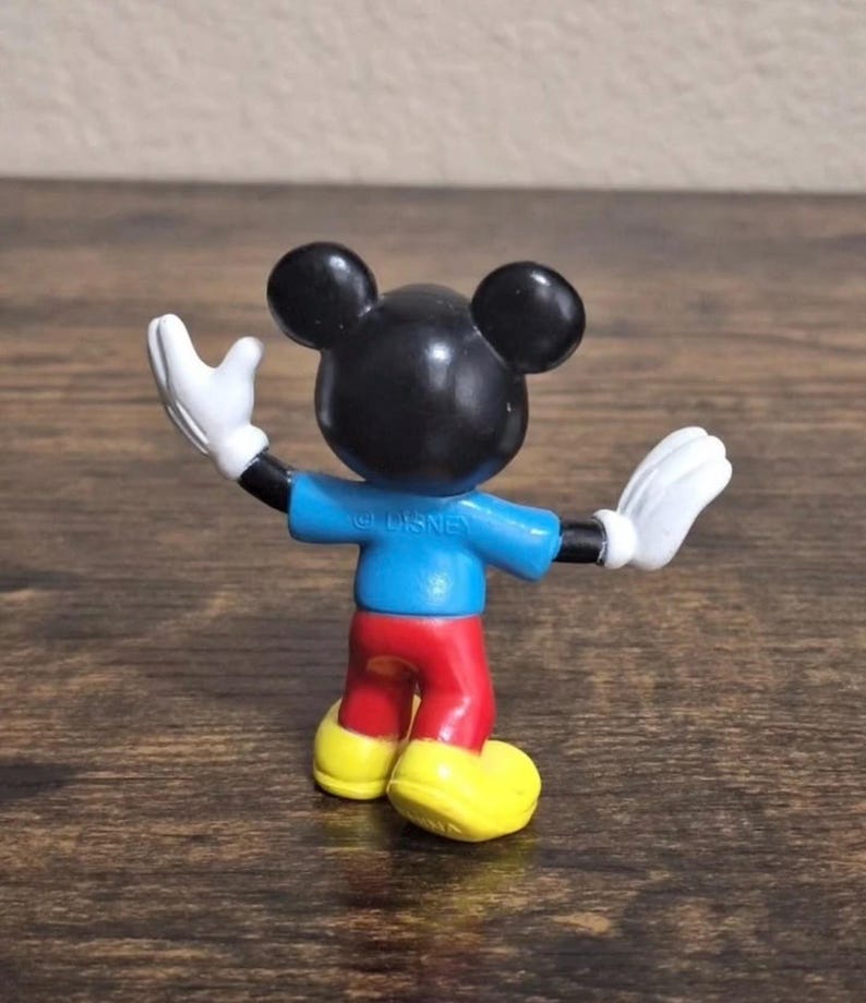 May include: A small Mickey Mouse figurine, viewed from the back, with arms outstretched. The figure has a black head with large ears, a blue shirt, red pants, yellow shoes, and white gloves. The word "DISNEY" is printed on the shirt.