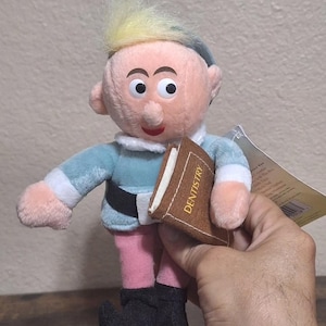 May include: A plush elf doll with blonde hair, wearing a blue and white outfit, holding a brown book with the word "Dentistry" printed on the cover.