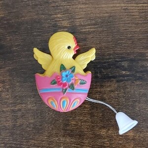 May include: A yellow chick with red beak and eyes peeks out of a pink and blue decorated egg with flowers. A white bell is attached to the egg with a string.