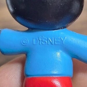 May include: Close-up of a Mickey Mouse figurine. The figure has a black head, blue shirt with the text "&copy; DISNEY", red shorts, and white gloves. The figure is a collectible item.