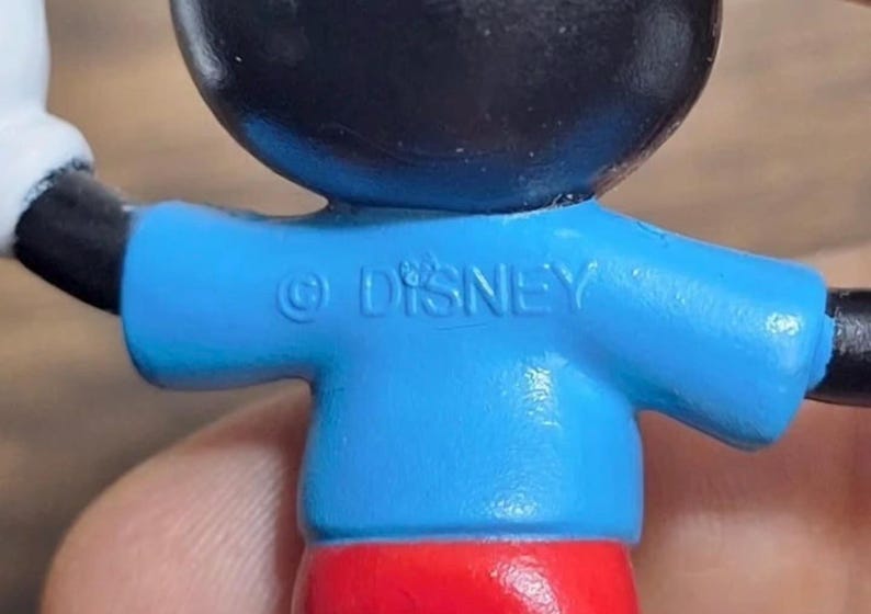 May include: Close-up of a Mickey Mouse figurine. The figure has a black head, blue shirt with the text "&copy; DISNEY", red shorts, and white gloves. The figure is a collectible item.