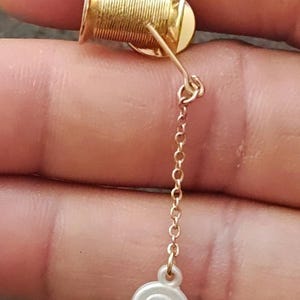 May include: A gold-colored earring featuring a miniature spool of thread with a dangling chain and a small, round, white charm. The charm has a floral design. The earring is a unique accessory.
