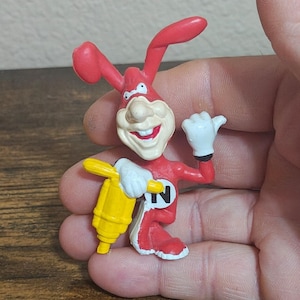 May include: A red and white cartoon rabbit figurine holding a yellow jackhammer. The rabbit has a large nose, a wide smile, and white gloves. The figurine has the letter "N" on its chest.
