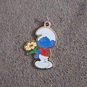 May include: A Smurf pendant with a gold-colored outline. The Smurf is blue with a white hat, red shirt, and white pants, holding a yellow flower with green leaves. The pendant has a small loop at the top for attachment.