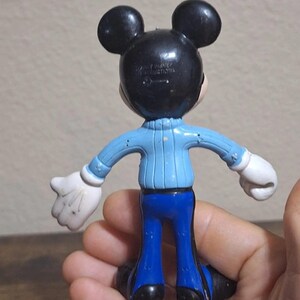 Puede incluir: Una figura vintage de Mickey Mouse, vista desde atr&aacute;s, con orejas y cabeza negras, un su&eacute;ter azul claro, pantalones azules, guantes blancos y zapatos negros. La figura se sostiene en una mano.