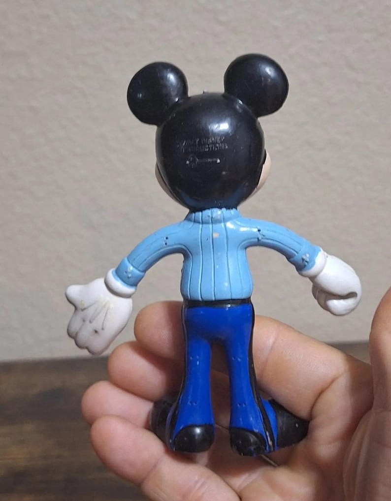 Puede incluir: Una figura vintage de Mickey Mouse, vista desde atr&aacute;s, con orejas y cabeza negras, un su&eacute;ter azul claro, pantalones azules, guantes blancos y zapatos negros. La figura se sostiene en una mano.