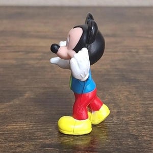 May include: A small Mickey Mouse figurine. The figure has a black head and ears, a pink face, white gloves, a blue shirt, red pants, and yellow shoes. The figurine is posed with its arms outstretched.