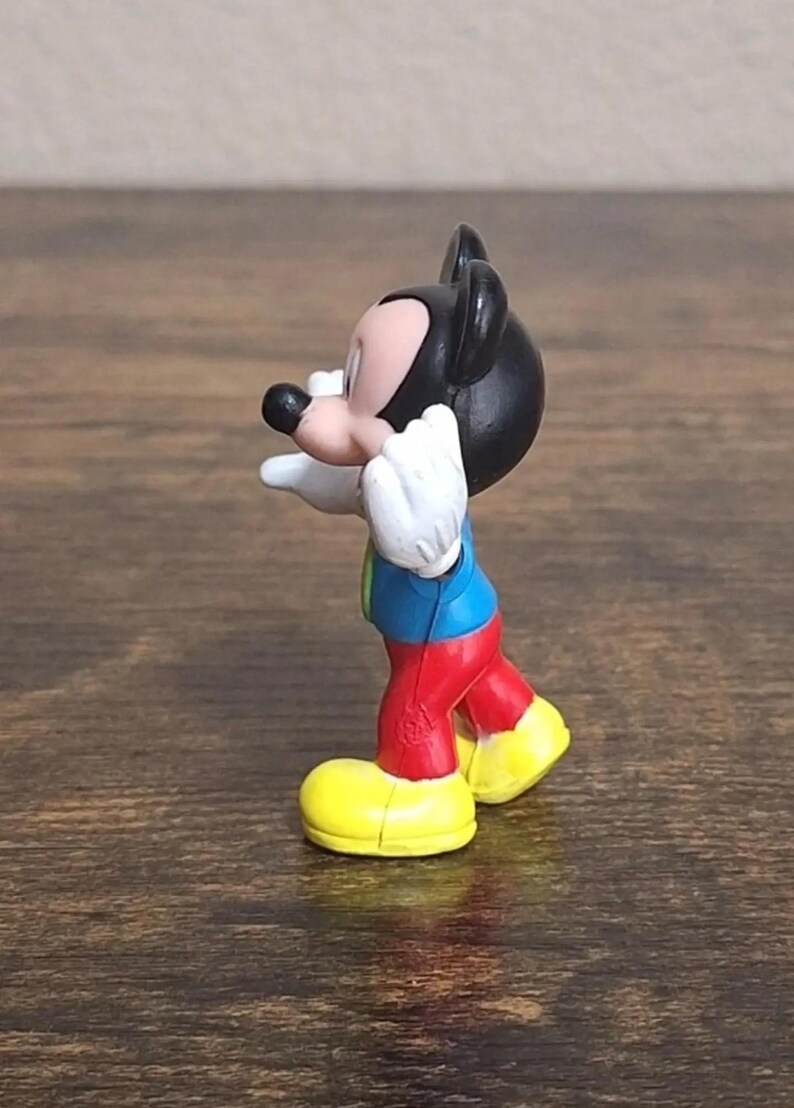 May include: A small Mickey Mouse figurine. The figure has a black head and ears, a pink face, white gloves, a blue shirt, red pants, and yellow shoes. The figurine is posed with its arms outstretched.