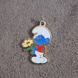May include: A gold-colored Smurf charm holding a yellow flower with green leaves. The Smurf is blue with a white hat, red shirt, and white pants. The charm has a small loop at the top for attaching to a chain or bracelet.
