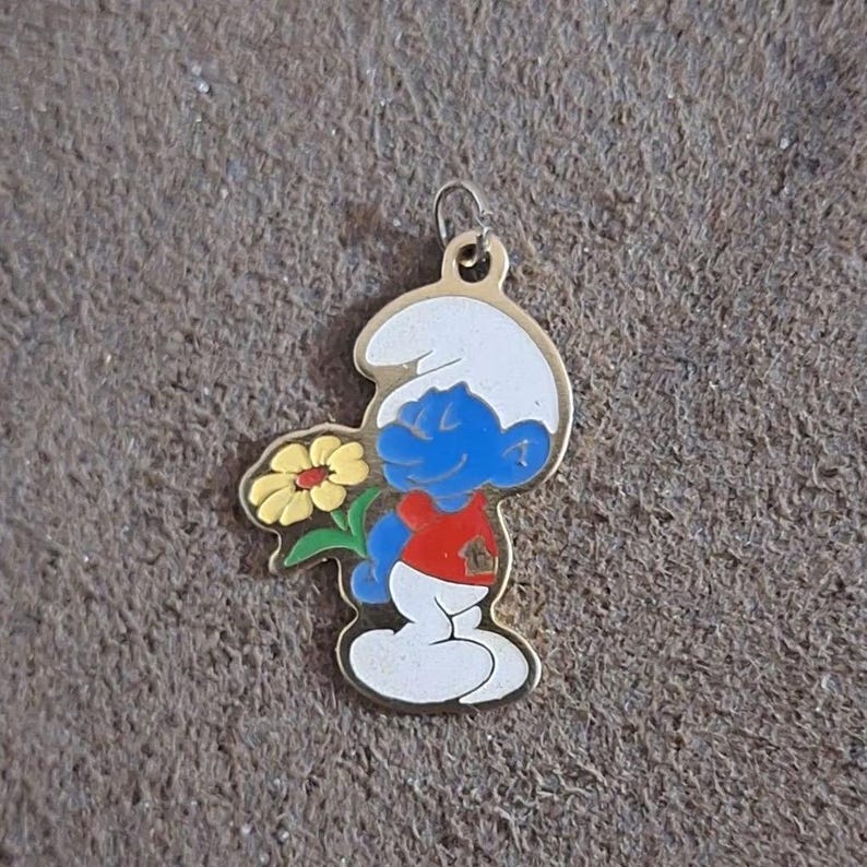 May include: A gold-colored Smurf charm holding a yellow flower with green leaves. The Smurf is blue with a white hat, red shirt, and white pants. The charm has a small loop at the top for attaching to a chain or bracelet.