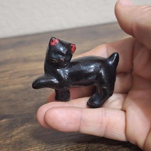 May include: A small, glossy black cat figurine with red accents on the eyes and nose. The cat is posed with one paw raised, giving it a playful appearance. The figurine is held in a hand.