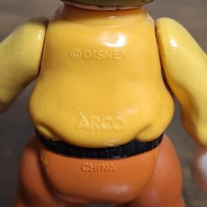 Puede incluir: Una figura de juguete vintage con torso amarillo, pantalones naranjas y cintur&oacute;n negro. La figura tiene las inscripciones "&copy;DISNEY", "ARGO" y "CHINA". Los brazos del juguete son amarillos y la cabeza es negra.