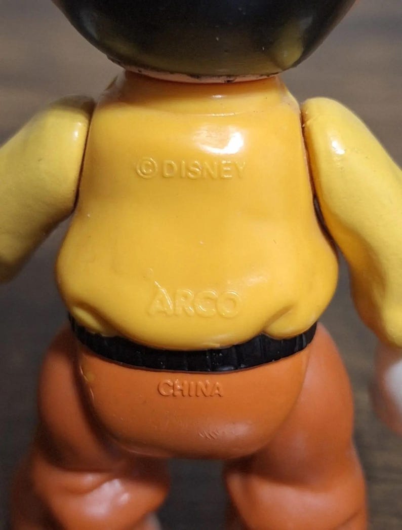 Puede incluir: Una figura de juguete vintage con torso amarillo, pantalones naranjas y cintur&oacute;n negro. La figura tiene las inscripciones "&copy;DISNEY", "ARGO" y "CHINA". Los brazos del juguete son amarillos y la cabeza es negra.