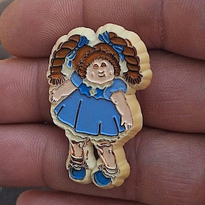 May include: A plastic pin featuring a cartoon doll with brown pigtails, a blue dress, and a white background.