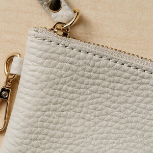 K&ouml;nnte beinhalten: Nahaufnahme einer hellgrauen Ledertasche mit goldenem Rei&szlig;verschluss und Beschl&auml;gen. Die Tasche hat eine Handschlaufe, die mit einem goldenen Verschluss befestigt ist. Das Leder hat ein strukturiertes, genarbtes Aussehen.