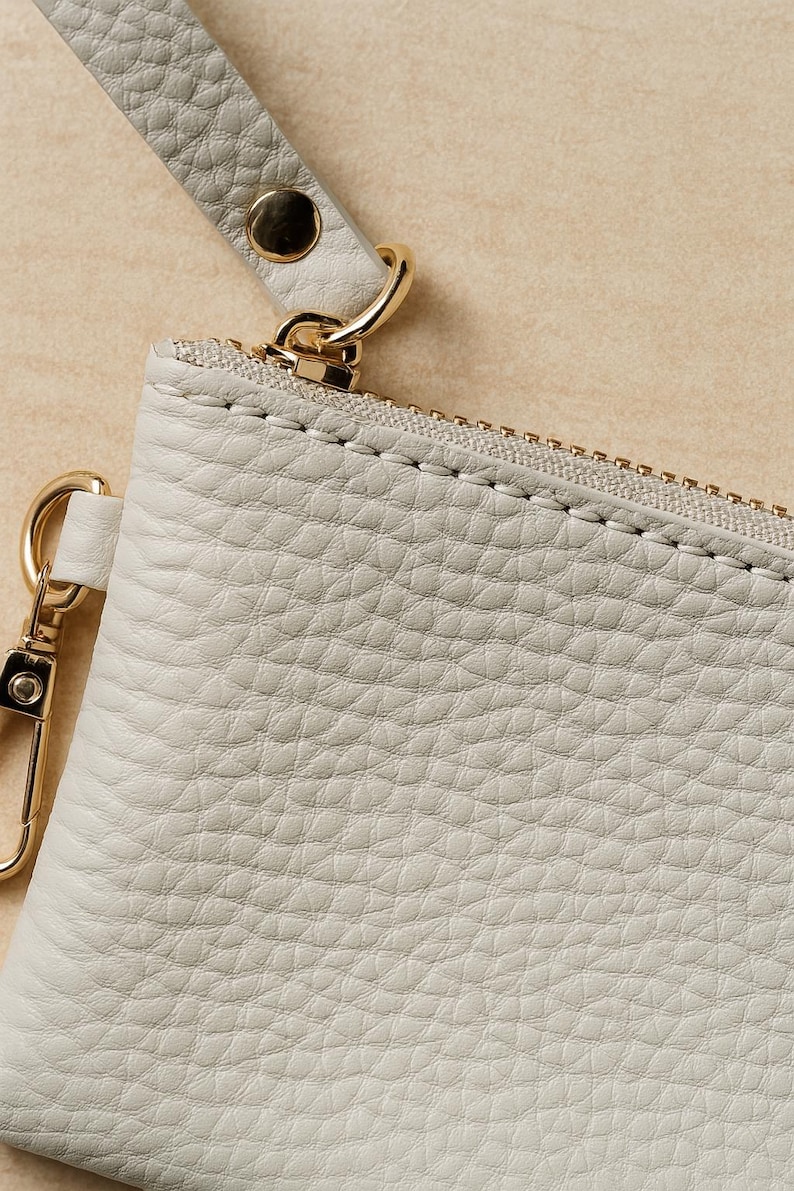K&ouml;nnte beinhalten: Nahaufnahme einer hellgrauen Ledertasche mit goldenem Rei&szlig;verschluss und Beschl&auml;gen. Die Tasche hat eine Handschlaufe, die mit einem goldenen Verschluss befestigt ist. Das Leder hat ein strukturiertes, genarbtes Aussehen.