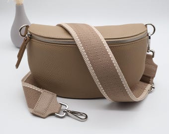 May include: A tan-colored crossbody bag with a textured leather appearance. The bag features a wide, patterned strap in beige and cream, and a silver zipper. The bag is designed with a curved shape and includes a small, matching leather pull.