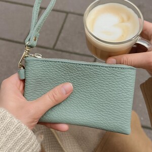 K&ouml;nnte beinhalten: Eine hellgr&uuml;ne Leder-Handgelenktasche mit Rei&szlig;verschluss, 12 cm x 7,5 cm gro&szlig;, wird von einer Person gehalten. Ein klares Glas Kaffee mit schaumiger Oberfl&auml;che wird in der N&auml;he gehalten. Die Handgelenktasche hat einen passenden Riemen und goldfarbene Hardware.