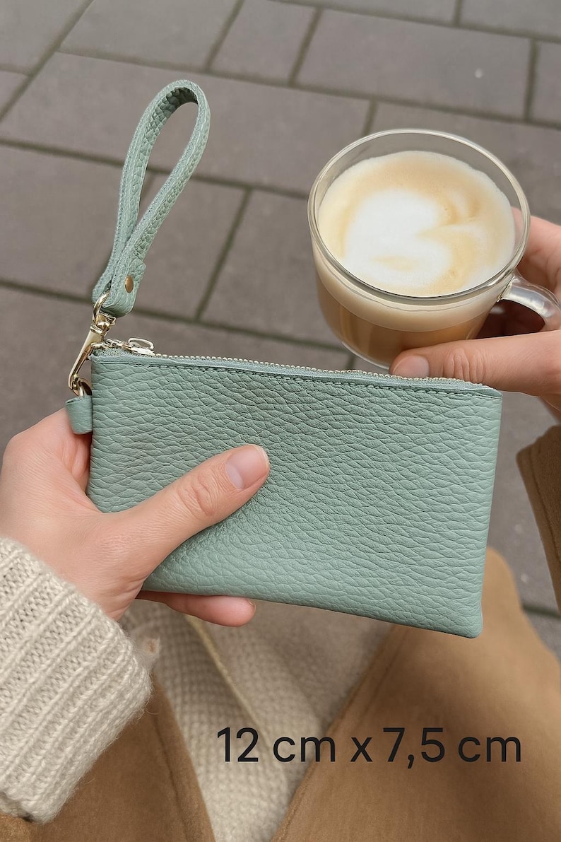 K&ouml;nnte beinhalten: Eine hellgr&uuml;ne Leder-Handgelenktasche mit Rei&szlig;verschluss, 12 cm x 7,5 cm gro&szlig;, wird von einer Person gehalten. Ein klares Glas Kaffee mit schaumiger Oberfl&auml;che wird in der N&auml;he gehalten. Die Handgelenktasche hat einen passenden Riemen und goldfarbene Hardware.