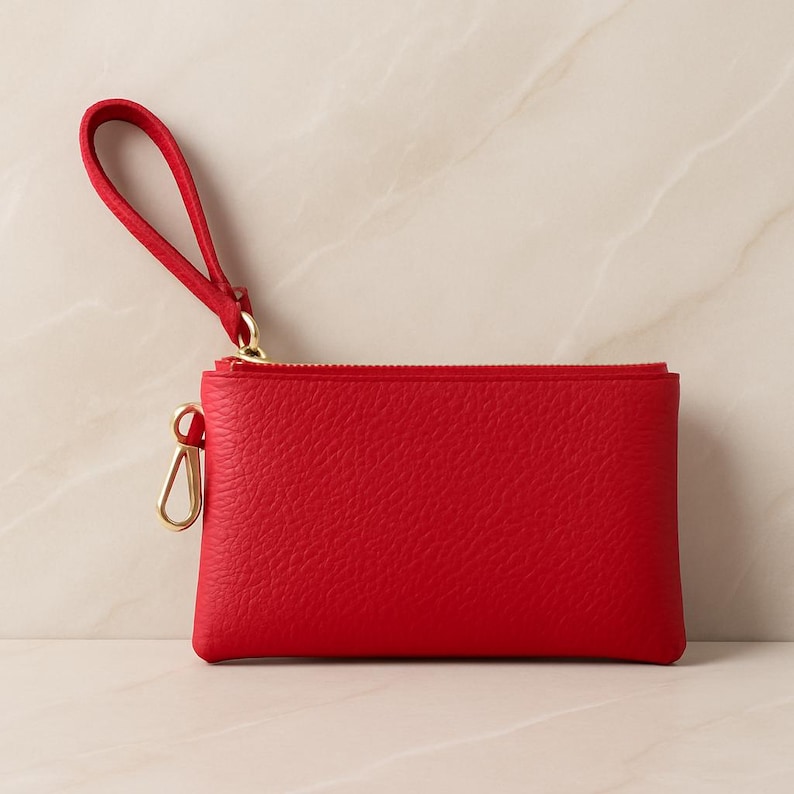 K&ouml;nnte beinhalten: Eine leuchtend rote Handgelenktasche mit goldenem Rei&szlig;verschluss und Beschl&auml;gen. Die kleine, rechteckige Geldb&ouml;rse hat eine Handschlaufe und eine strukturierte Oberfl&auml;che. Die Handgelenktasche ist f&uuml;r den Transport kleiner Essentials konzipiert.