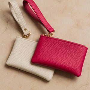 K&ouml;nnte beinhalten: Zwei kleine Leder-M&uuml;nzb&ouml;rsen. Eine ist leuchtend pink, die andere cremefarben. Beide haben eine Handschlaufe und einen goldfarbenen Rei&szlig;verschluss und Beschl&auml;ge. Die Geldb&ouml;rsen sind aus strukturiertem Leder gefertigt und werden auf einer neutralen Oberfl&auml;che pr&auml;sentiert.