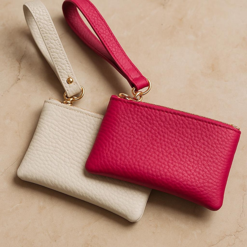 K&ouml;nnte beinhalten: Zwei kleine Leder-M&uuml;nzb&ouml;rsen. Eine ist leuchtend pink, die andere cremefarben. Beide haben eine Handschlaufe und einen goldfarbenen Rei&szlig;verschluss und Beschl&auml;ge. Die Geldb&ouml;rsen sind aus strukturiertem Leder gefertigt und werden auf einer neutralen Oberfl&auml;che pr&auml;sentiert.