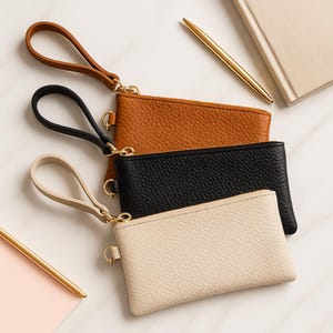K&ouml;nnte beinhalten: Eine Sammlung von drei Leder-Handgelenktaschen in den Farben Tan, Schwarz und Creme. Jede Geldb&ouml;rse hat eine passende Handschlaufe und goldfarbene Hardware. Die Geldb&ouml;rsen liegen auf einer wei&szlig;en Marmoroberfl&auml;che, mit einem goldenen Stift und Notizbuch im Hintergrund.