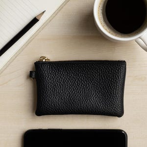 K&ouml;nnte beinhalten: Eine schwarze Leder-Geldb&ouml;rse mit goldenem Rei&szlig;verschluss, ein Smartphone, eine Tasse Kaffee, ein Notizblock und ein Bleistift sind auf einer hellen Holzoberfl&auml;che angeordnet. Die Geldb&ouml;rse hat eine strukturierte Oberfl&auml;che.