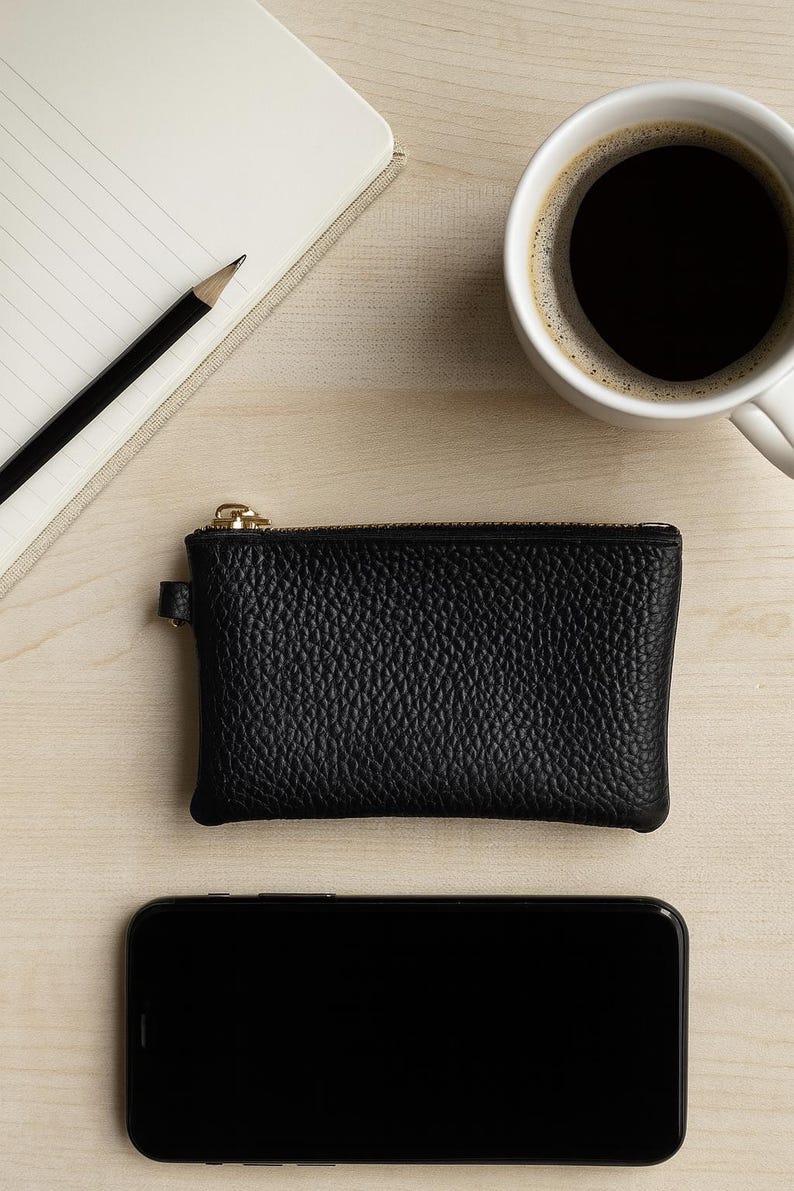 K&ouml;nnte beinhalten: Eine schwarze Leder-Geldb&ouml;rse mit goldenem Rei&szlig;verschluss, ein Smartphone, eine Tasse Kaffee, ein Notizblock und ein Bleistift sind auf einer hellen Holzoberfl&auml;che angeordnet. Die Geldb&ouml;rse hat eine strukturierte Oberfl&auml;che.