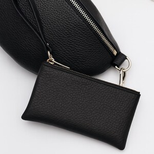 K&ouml;nnte beinhalten: Schwarze Leder-Bauchtasche mit passendem kleinem Handgelenkbeutel. Beide Artikel haben eine strukturierte Oberfl&auml;che und silberfarbene Rei&szlig;verschl&uuml;sse. Die Bauchtasche hat eine Handschlaufe und einen silberfarbenen Ring.