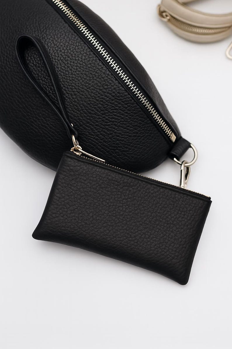 K&ouml;nnte beinhalten: Schwarze Leder-Bauchtasche mit passendem kleinem Handgelenkbeutel. Beide Artikel haben eine strukturierte Oberfl&auml;che und silberfarbene Rei&szlig;verschl&uuml;sse. Die Bauchtasche hat eine Handschlaufe und einen silberfarbenen Ring.