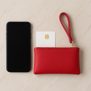 K&ouml;nnte beinhalten: Eine rote Leder-Handgelenktasche mit goldenem Rei&szlig;verschluss und passendem Handgelenkriemen. Eine wei&szlig;e Karte mit goldenem Chip ist teilweise in der Handgelenktasche sichtbar. Links von der Handgelenktasche befindet sich ein schwarzes Smartphone.