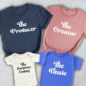 May include: Four matching t-shirts and a baby onesie. The shirts are navy, dusty rose, and blue, with the words "The Producer", "The Creator", and "The Finale" printed in white. The onesie is cream-colored and says "The Surprise Ending".