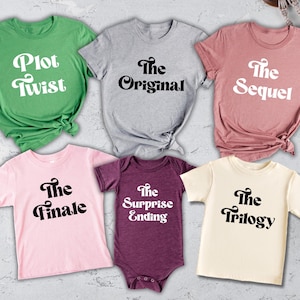 May include: A collection of t-shirts and a onesie in various colors, each featuring a different phrase in a retro font. The shirts include "Plot Twist," "The Original," "The Sequel," "The Finale," and "The Trilogy." The onesie says "The Surprise Ending."