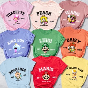 May include: A collection of colorful t-shirts featuring various characters from the Super Mario video game series. Each shirt displays a character's name, the year 1985, and the Super Mario logo. Colors include pink, peach, purple, green, orange, red, light blue, and yellow.