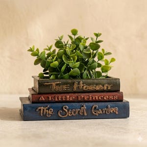 May include: A decorative planter shaped like a stack of books, with the titles "The Hobbit", "A Little Princess", and "The Secret Garden" embossed on the spines. The planter is filled with green leafy plants. The books are painted in shades of green, red, and blue.