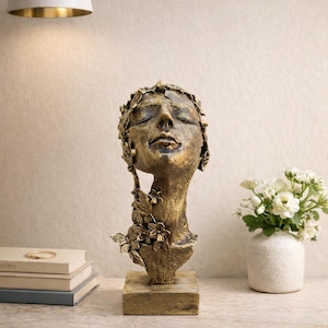 Könnte beinhalten: Eine bronzefarbene Skulptur eines Frauenkopfes mit geschlossenen Augen, verziert mit floralen Details. Die Skulptur steht auf einem quadratischen Sockel. Eine Vase mit weißen Blumen und ein Stapel Bücher befinden sich in der Nähe.