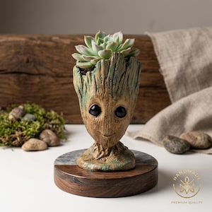 Pode incluir: Um vaso em forma de personagem de árvore castanho e verde com grandes olhos pretos, contendo uma planta suculenta verde. O vaso assenta numa base circular de madeira com um topo de mármore e madeira. O texto "HANDMADE PREMIUM QUALITY" é visível.