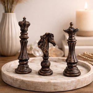May include: Three bronze chess pieces: a king, a knight, and a queen, displayed on a marble tray. The pieces have intricate details and a vintage aesthetic. A lit candle and decorative vase are in the background.