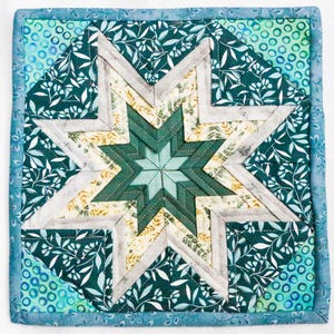 Puede incluir: Textil acolchado cuadrado con un diseño de estrella central. La estrella está compuesta por varios tonos de verde, gris y amarillo. El fondo es de tela verde azulado con un estampado de hojas blancas. El borde es de tela verde azulado con un estampado de puntos azules y verdes.