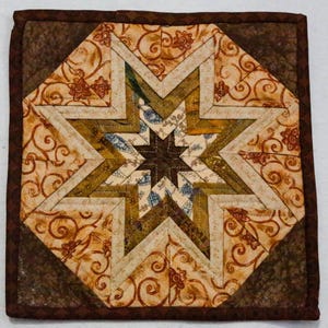 Puede incluir: Un posavasos cuadrado acolchado con un diseño de estrella. La estrella está hecha de telas en tonos marrón, beige y verde oliva. El borde exterior es de color marrón oscuro. El artículo hecho a mano tiene una estética rústica y cálida.