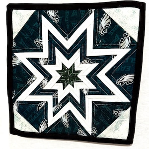 Könnte beinhalten: Ein quadratischer, gesteppter Untersetzer mit einem Sternmuster. Die Mitte ist dunkelgrün, umgeben von weißem und dunkelgrünem Stoff mit dem Logo der Philadelphia Eagles. Die Kanten sind mit schwarzem Binding versehen.